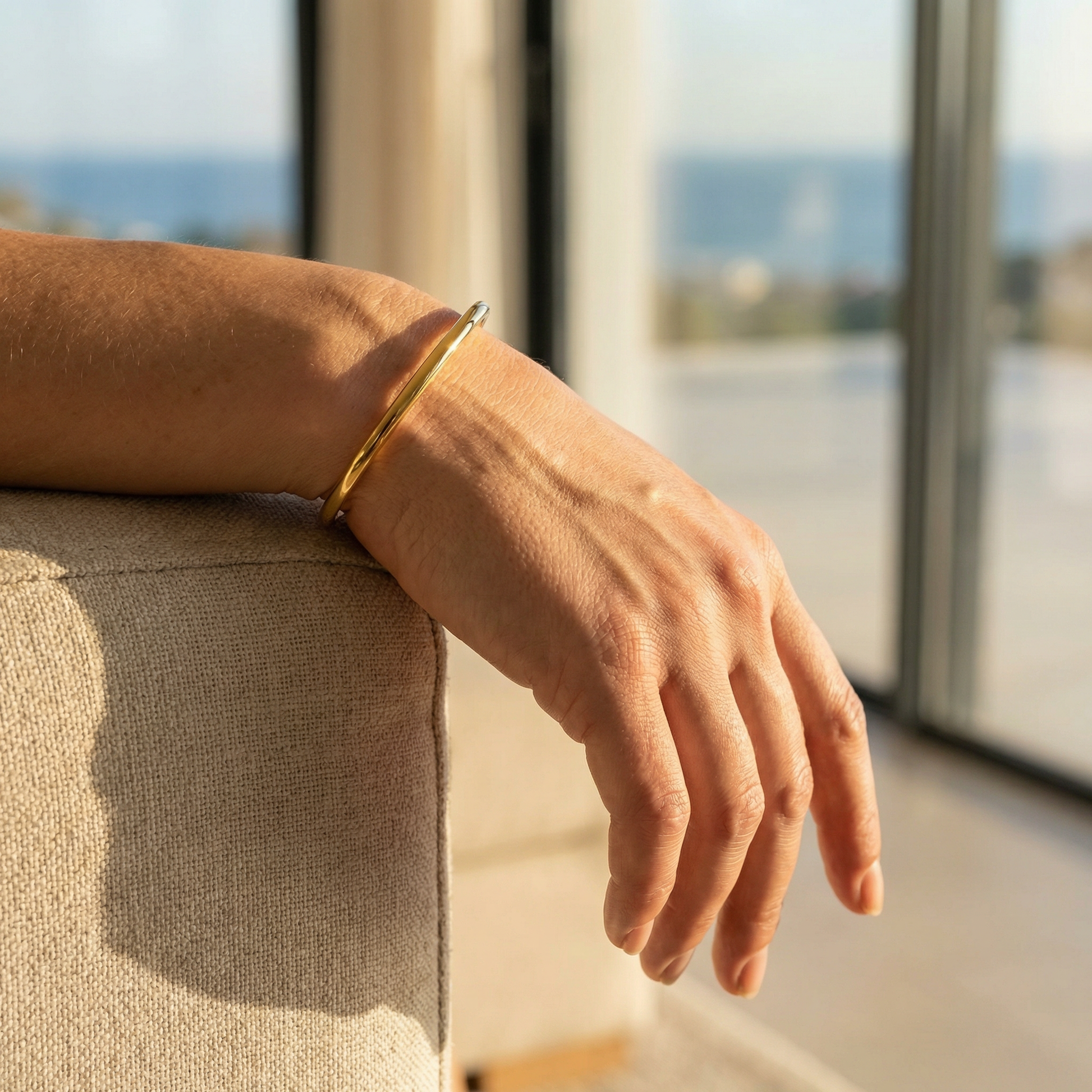 Hand wearing a gold bangle by a window with a view of water