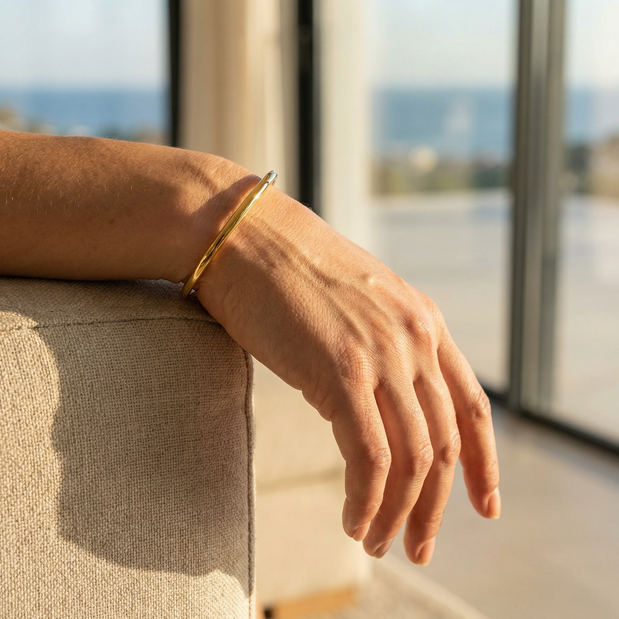 Hand wearing a gold bangle by a window with a view of water