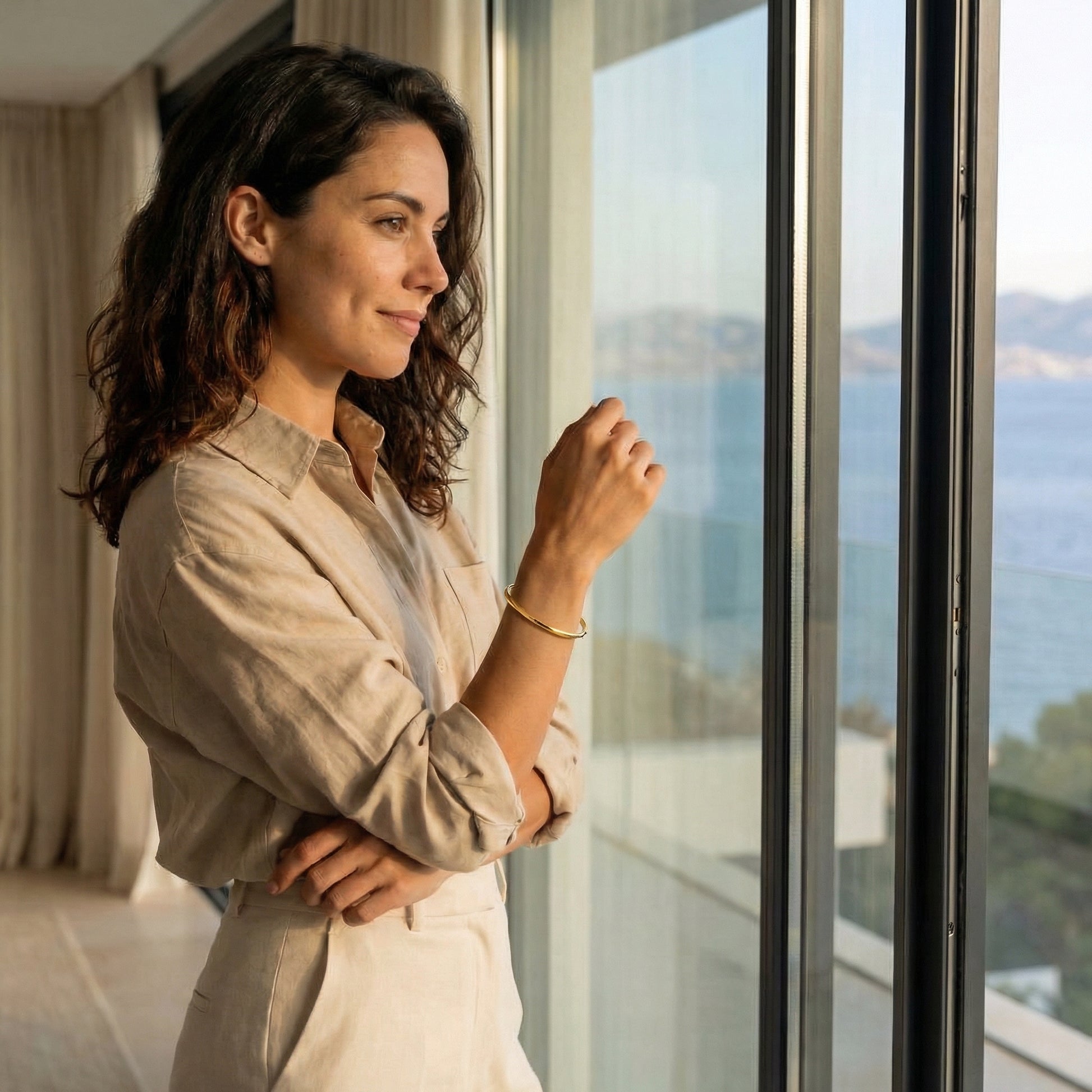 Woman standing by a window with a scenic view, wearing a beige shirt and bangle around the wrist.