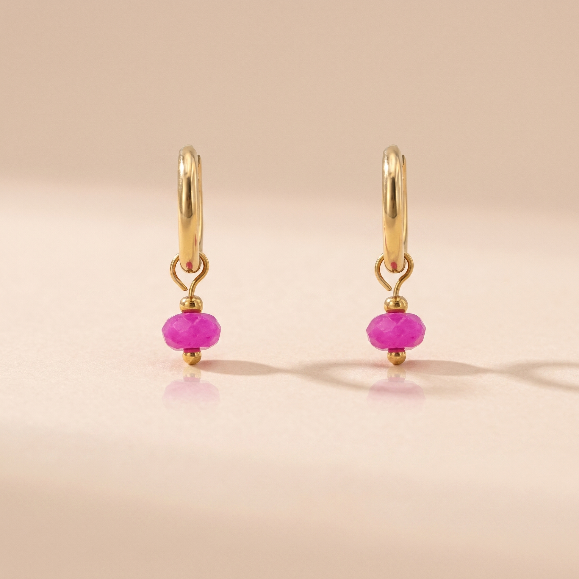Gold-tone huggie hoop earrings with fuchsia natural stone on a beige background