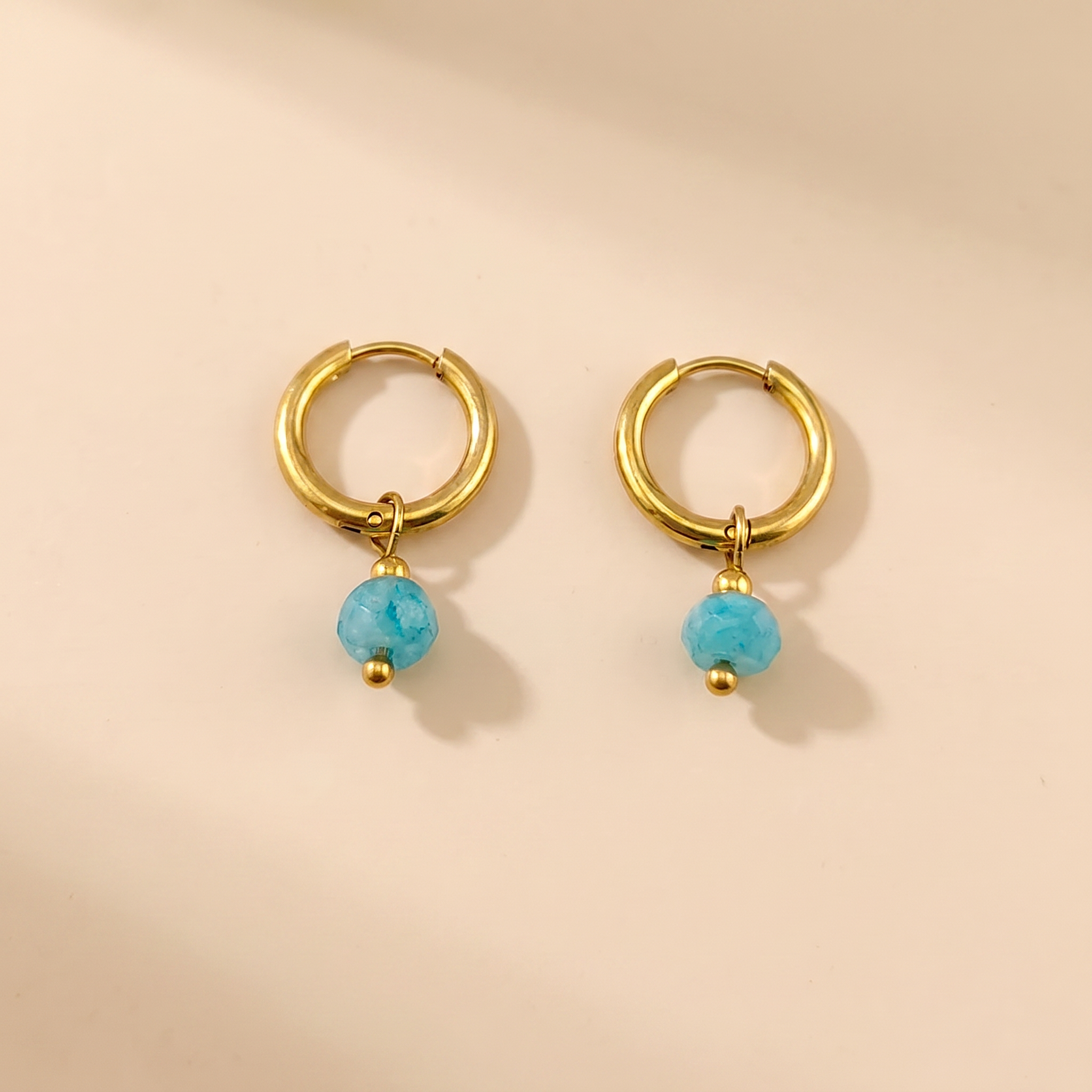 Gold-tone huggie hoop earrings with blue natural stone on a beige background