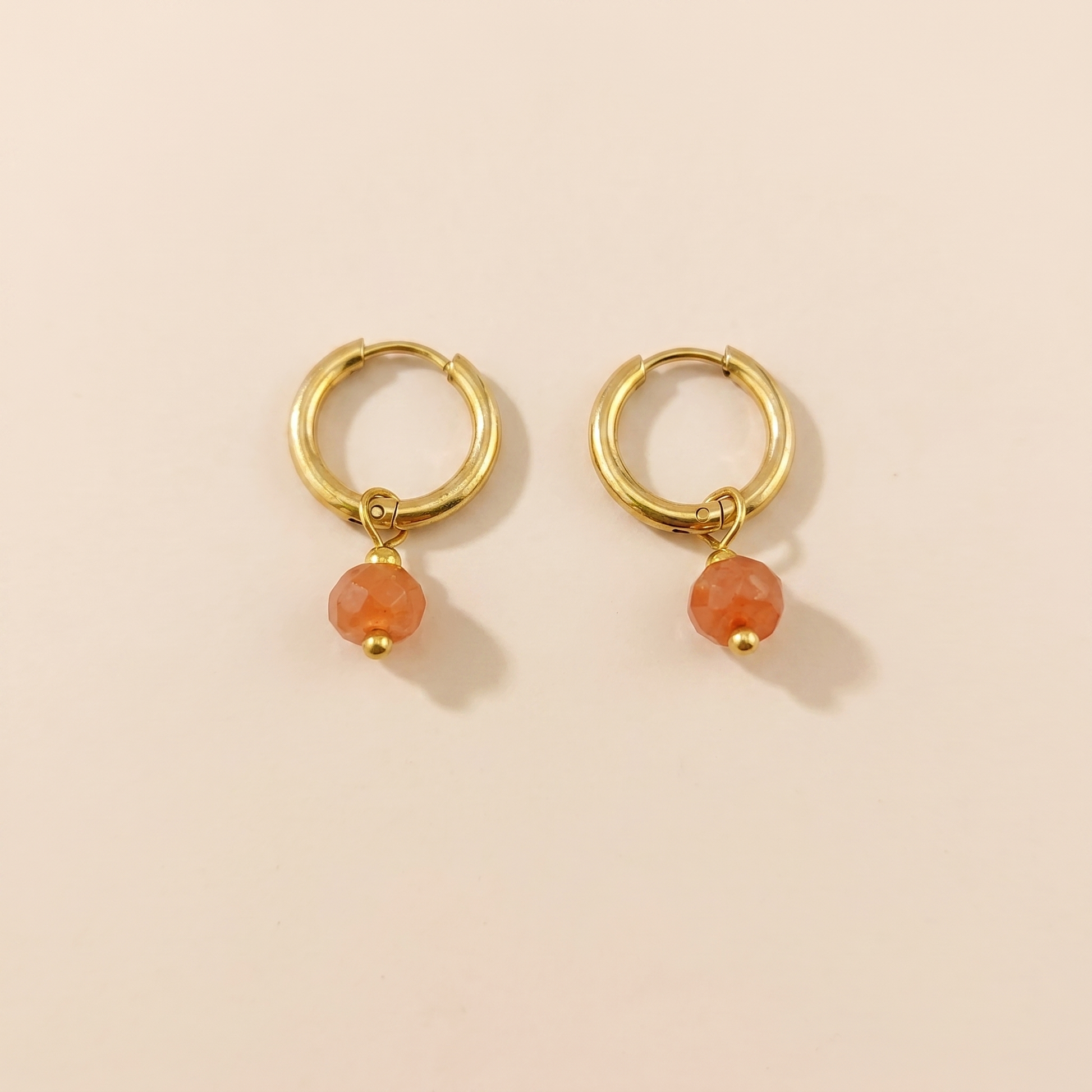 Gold-tone huggie hoop earrings with orange natural stone on a beige background