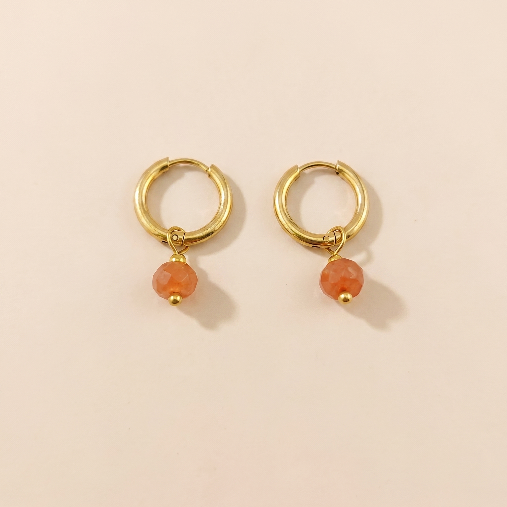 Gold-tone huggie hoop earrings with orange natural stone on a beige background